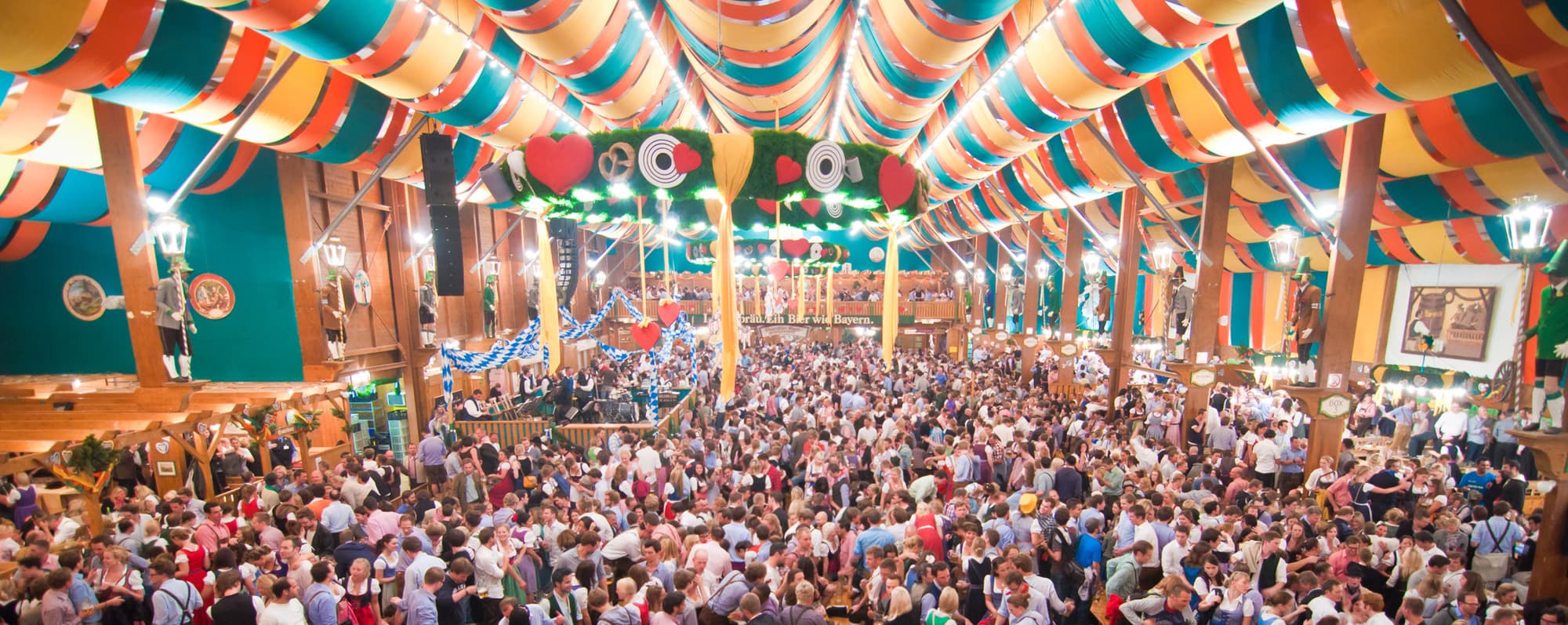 Interiör från öltält på Oktoberfest. Längs långa bord sitter människor som äter och dricker.